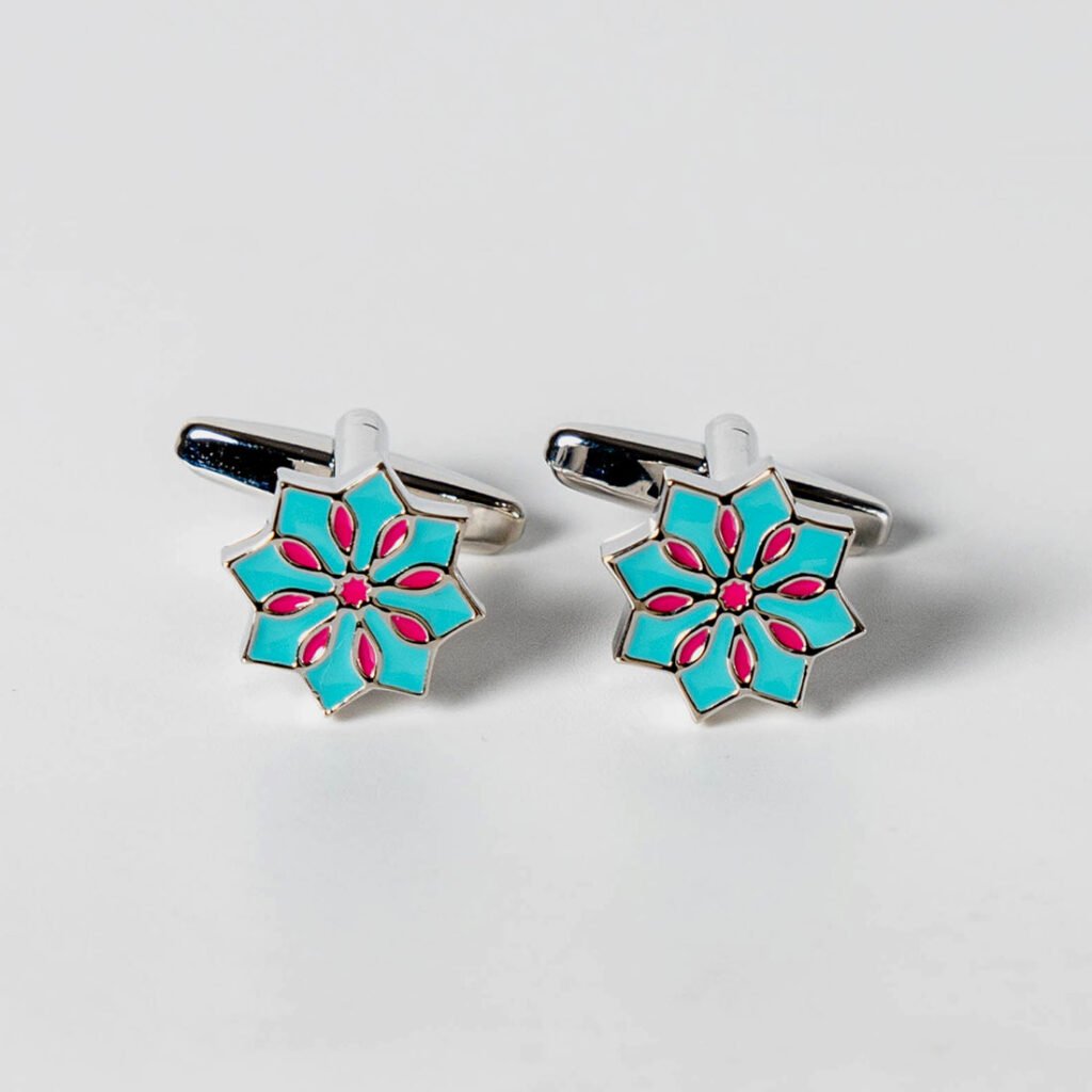 A pair of silver-toned cufflinks with an eight-pointed star design. The star is filled with a vibrant teal color, and within it, a smaller eight-pointed design is present in a bright pink hue. The cufflinks are positioned on a white background, slightly angled, showcasing their faces and the clasps. Ethnic Mosaic Cufflinks - Blue